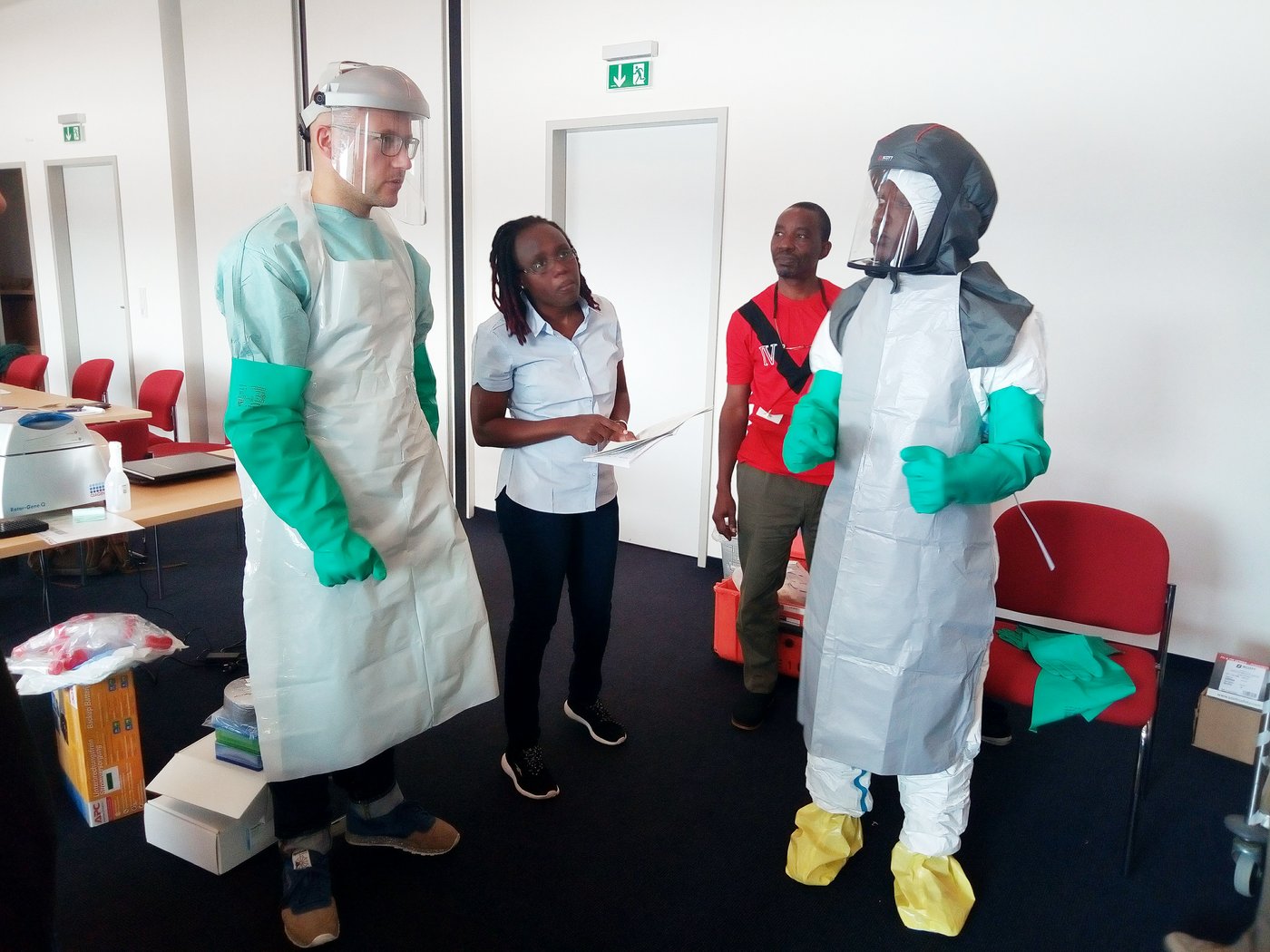 The picture shows a course situation with four scientists, two of them dressed in safety suits, standing together discussing.