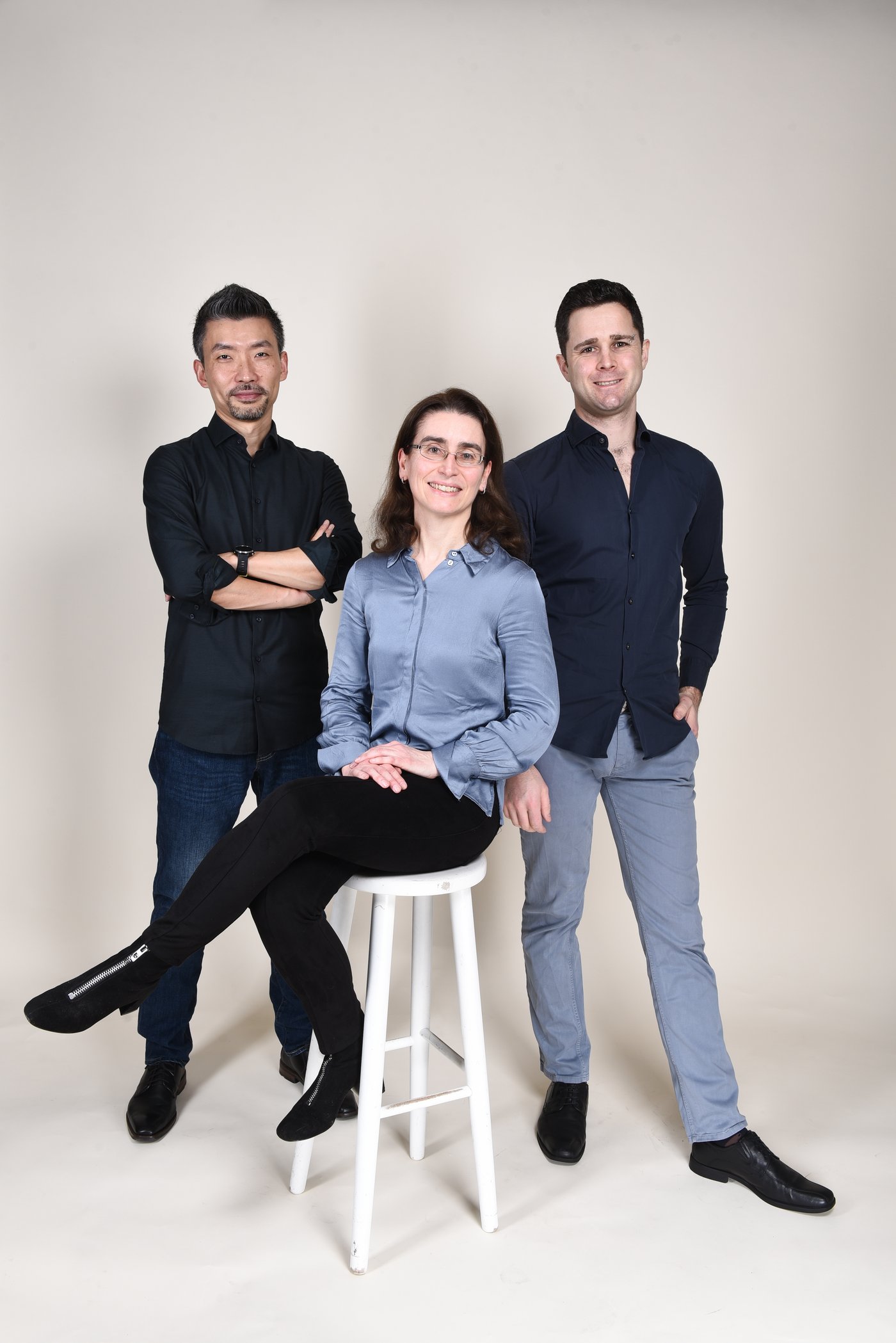 The picture of the Panadea team in front of a light beige background: A young woman with medium-length brown hair, a light blue blouse and black jeans is sitting on a white bar stool. Behind her are two young men in dark shirts and jeans.