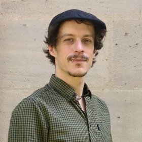 A man with curly hair and a mustache wearing a black flat cap and a green checkered shirt stands against a textured stone wall, looking at the camera.