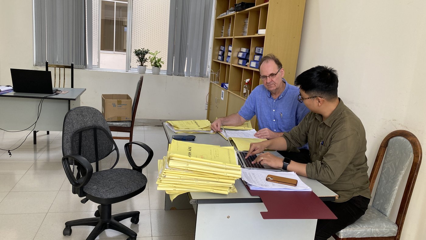 The picture shows two researchers in the office of a hospital. They are sitting at a table with many medical records, which they are looking at and discussing together. One researcher is typing the data on his laptop.
