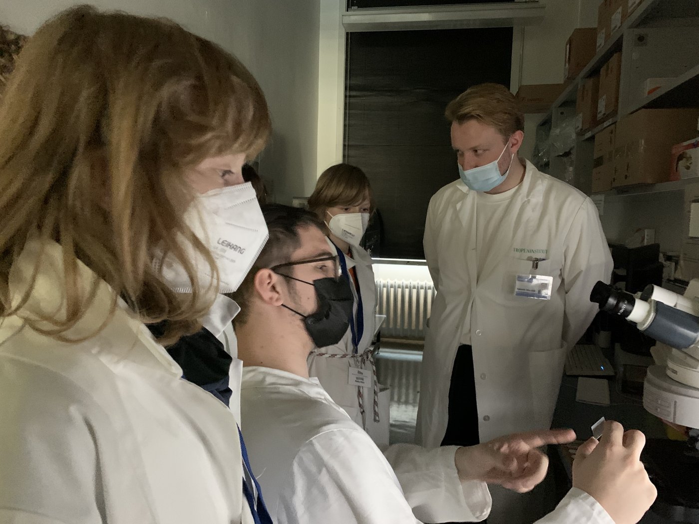 Mehrere Jungen schauen im Labor einem Forscher über die Schulter. Der Forscher sitzt vor einem Mikroskop und erklärt etwas.