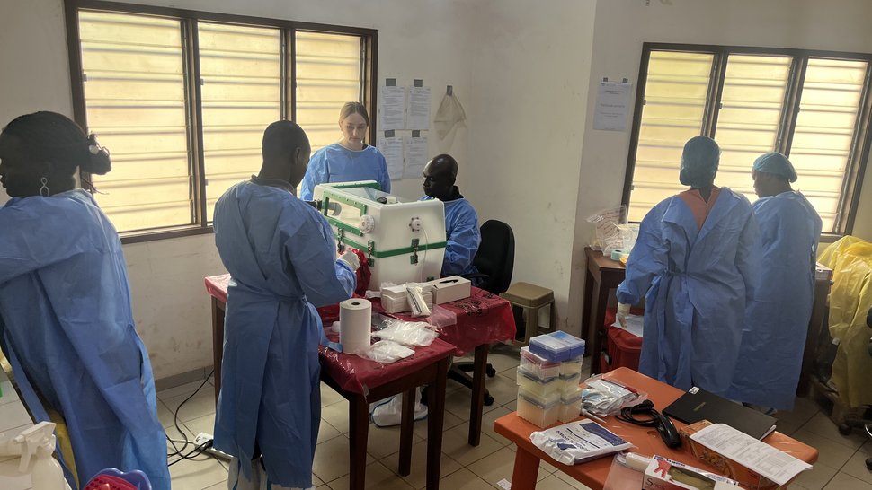 Laboratory Training for Diagnostic of Viral hemorrhgagic fever viruses at CIPEC-BA, Parakou, Benin Photo of several international scientists in a room with louvred windows. Two colleagues are working on the glove box.