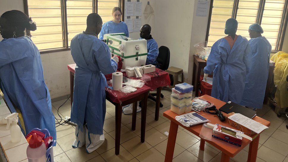 Foto mehrerer internationaler Wissenschaftler:innen in einem Raum mit Lamellenfenstern. Zwei Kollegen arbeiten an der Glove box.