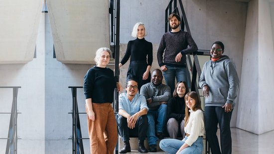 A group photo picturing eight diverse people in front of a staircase. Four are sitting in the front, surrounded by the others standing.