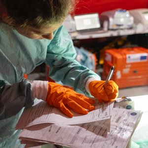 Das Foto zeigt eine Expertin in grünlichem Laborkittel und mit orangen Laborhandschuhen beim Notizen machen. Im Hintergrund orange Mobile-Lab-Kisten.