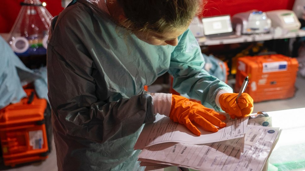Das Foto zeigt eine Expertin in grünlichem Laborkittel und mit orangen Laborhandschuhen beim Notizen machen. Im Hintergrund orange Mobile-Lab-Kisten.