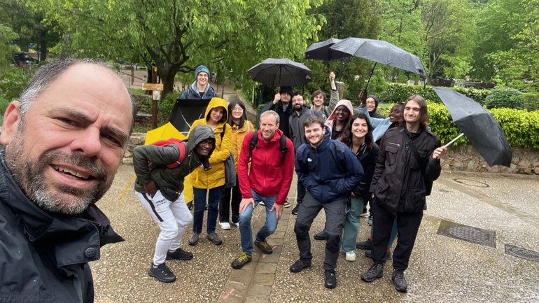 A group of people poses outdoors on a rainy day, some holding umbrellas and wearing rain jackets, with trees and greenery in the background.