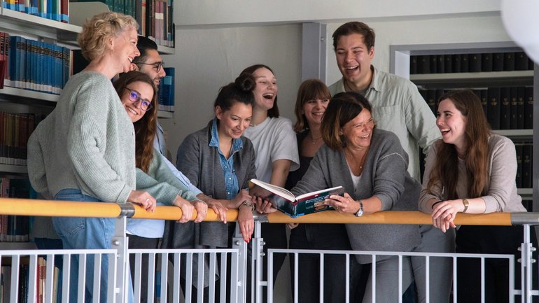 The working group stands at the railing of the gallery in the library. The working group leader is holding a book in her hand and laughing. Some of the other colleagues look over at her and laugh too.