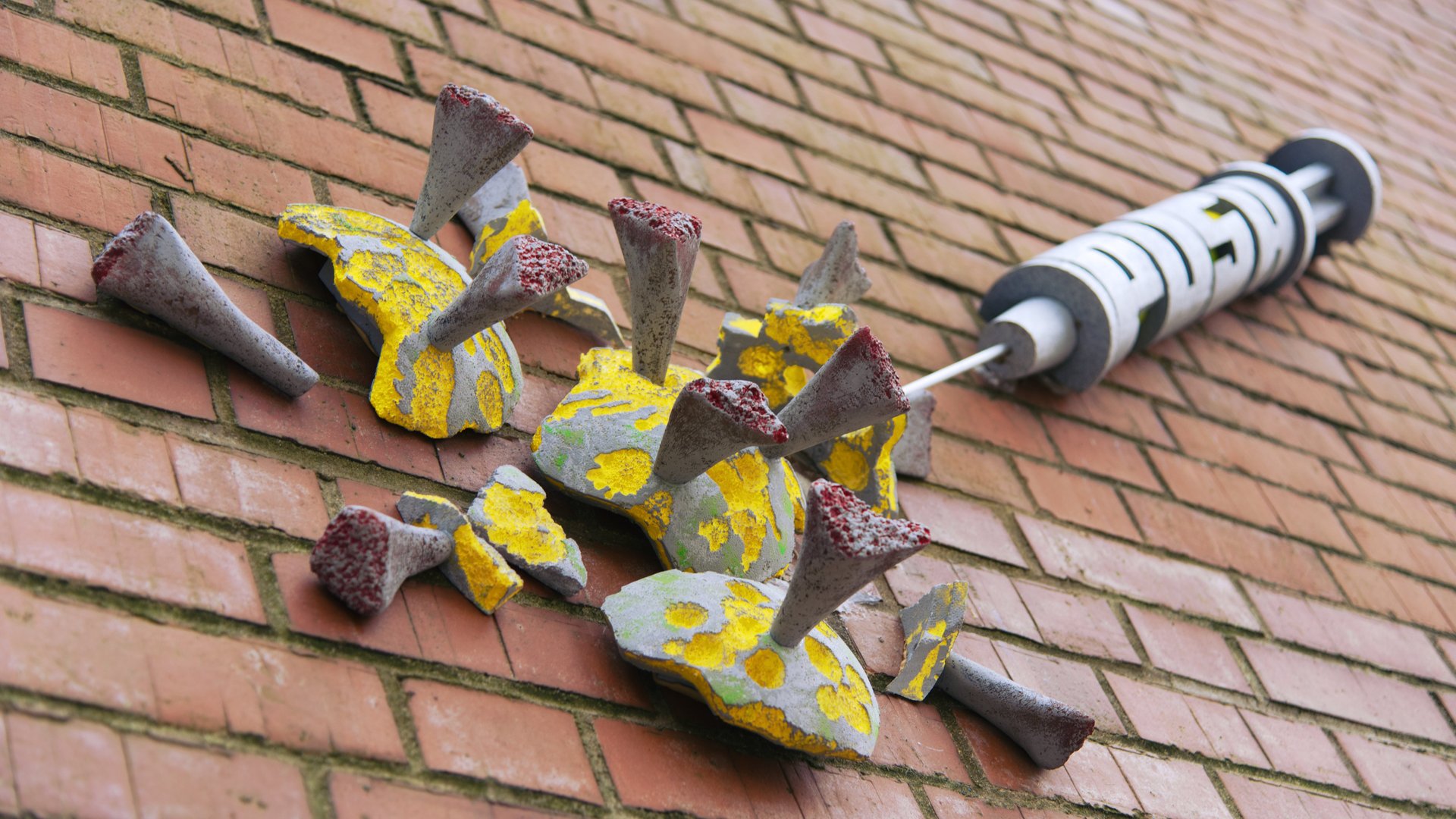 The photo shows a street art sculpture on a red brick wall: a yellow-grey-red SARS CoV-2 virus. A syringe with the inscription "Push" is coming from the top right. The virus appears to be bursting.