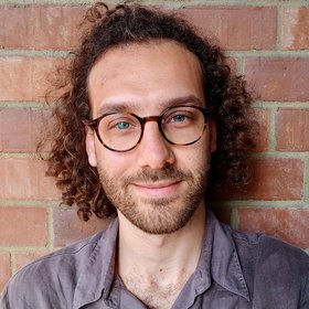 [Translate to English:] Gabriele Diana: A male person with curly brown, longer hair wearing glasses in front of a red brick wall is looking friendly into the camera.
