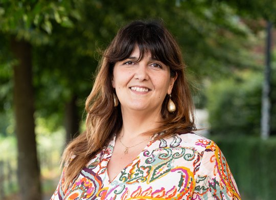 Das Foto zeigt eine Frau mit langen braunen Haaren. Sie trägt eine bunt gemusterte Bluse.