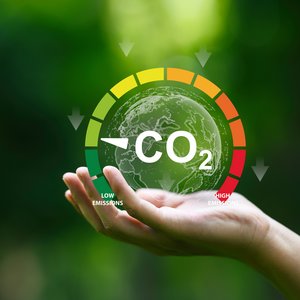 Den CO2-Ausstoß im Blick behalten Eine Hand hält einen leuchtenden Globus vor grünem, unscharfem Hintergrund. Darüber liegt eine Anzeige mit farbigem Bogen von Grün bis Rot. Der weiße Zeiger zeigt auf den grünen Bereich. In der Mitte steht „CO2“. Unten lauten die Beschriftungen „Low Emissions“ und „High Emissions“, also niedriger und hoher CO2-Ausstoß.