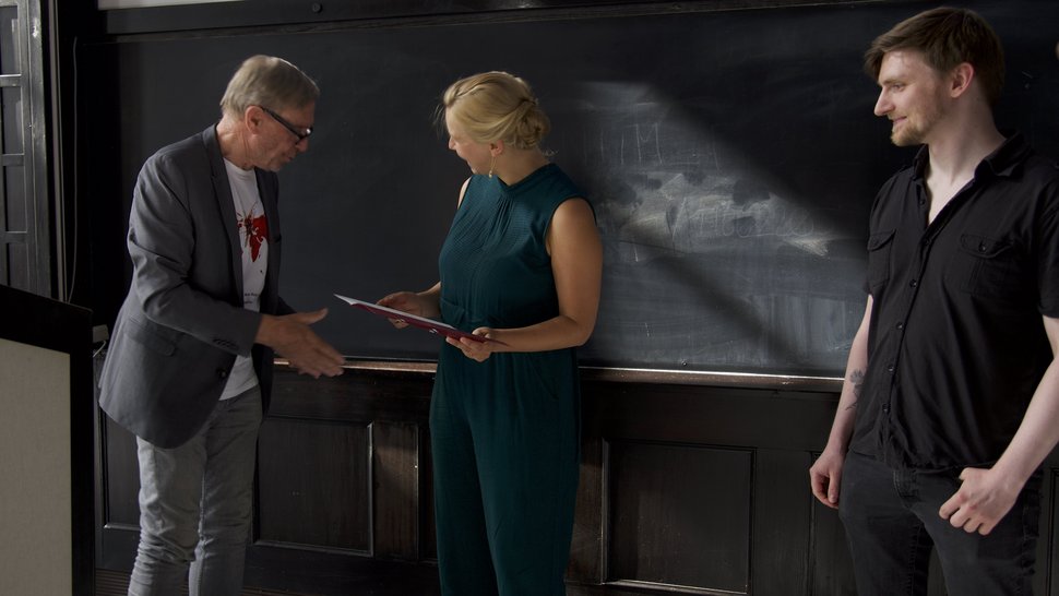 The photo shows three people at the award ceremony: The VdF Chairman on the left presents Wiebke Herr with her certificate. Further to the right, Chris Hoffmann is standing and smiling. In the background is the dark wood panelling of the historic lecture hall.