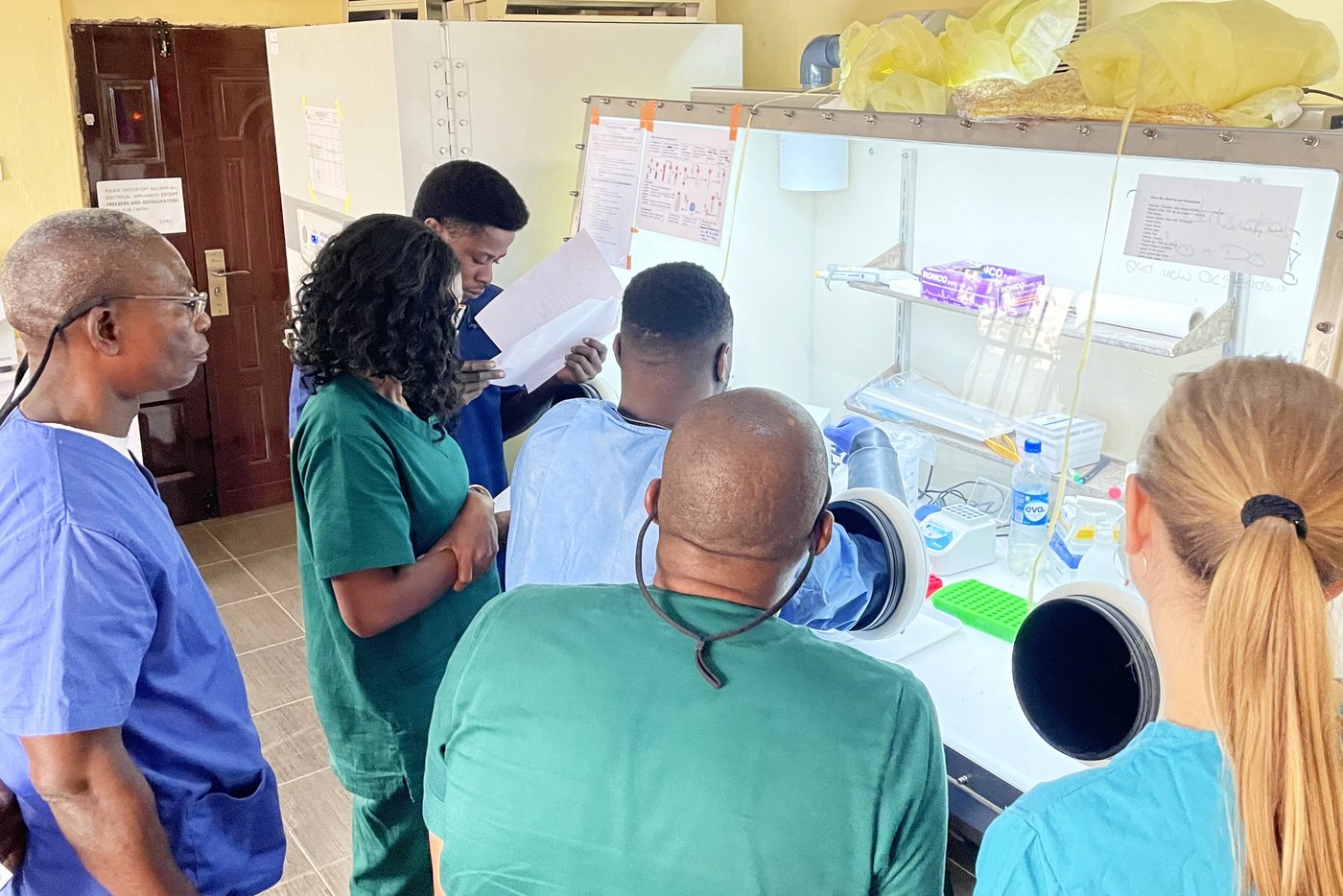Training of local scientists to safely inactivate infectious Lassa virus patient samples in a glovebox. Four laboratory trainees and one trainer stand around a glovebox during a hands-on training of one laboratory staff who is operating in a containment environment to learn how to safely inactivate infectious Lassa virus patient samples.