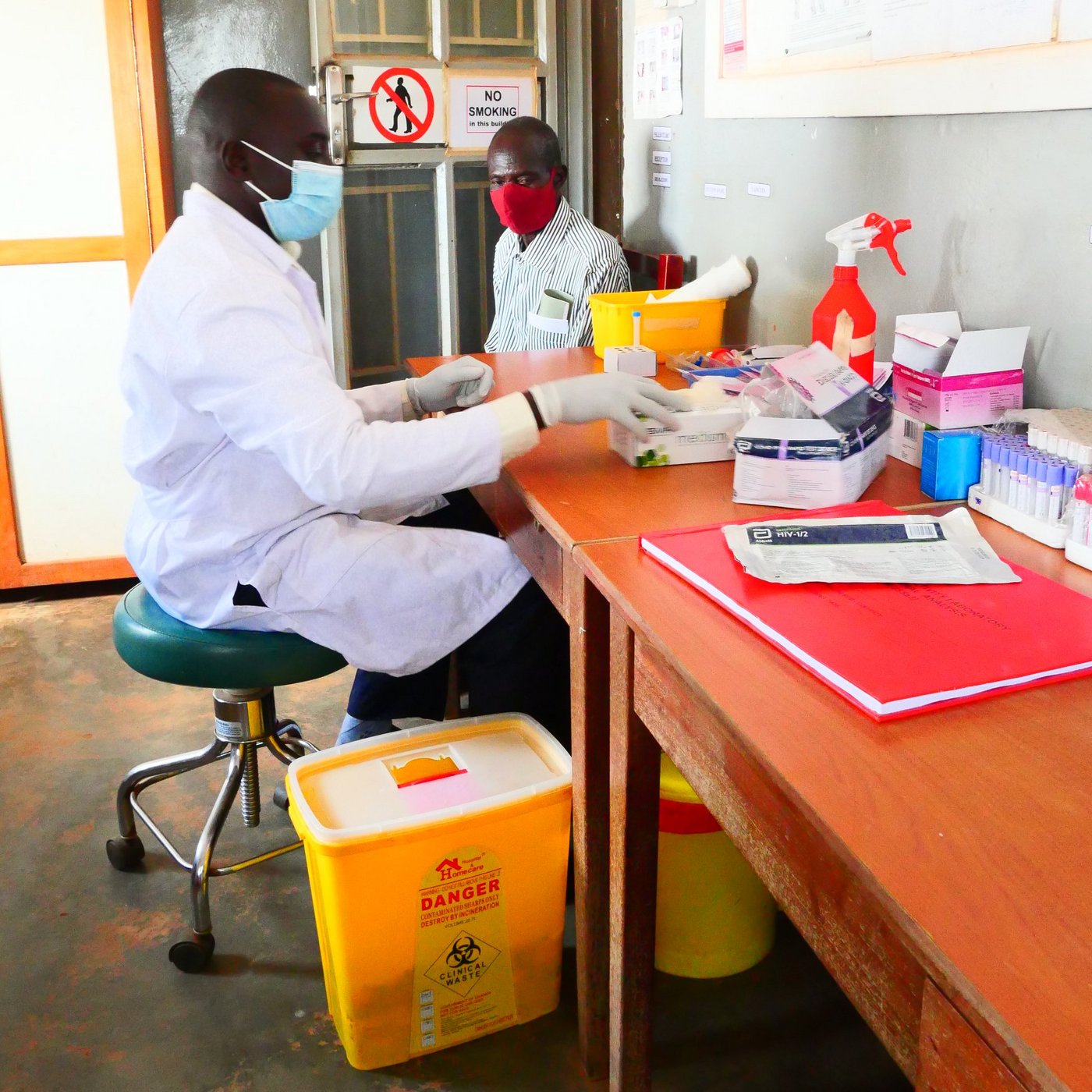 Covid 19 testing at Bobbi Hospital, Uganda. A person works at a table on which laboratory utensils are lying. The person is wearing gloves, a mask and a lab coat. Another person is sitting on a chair next to the table.