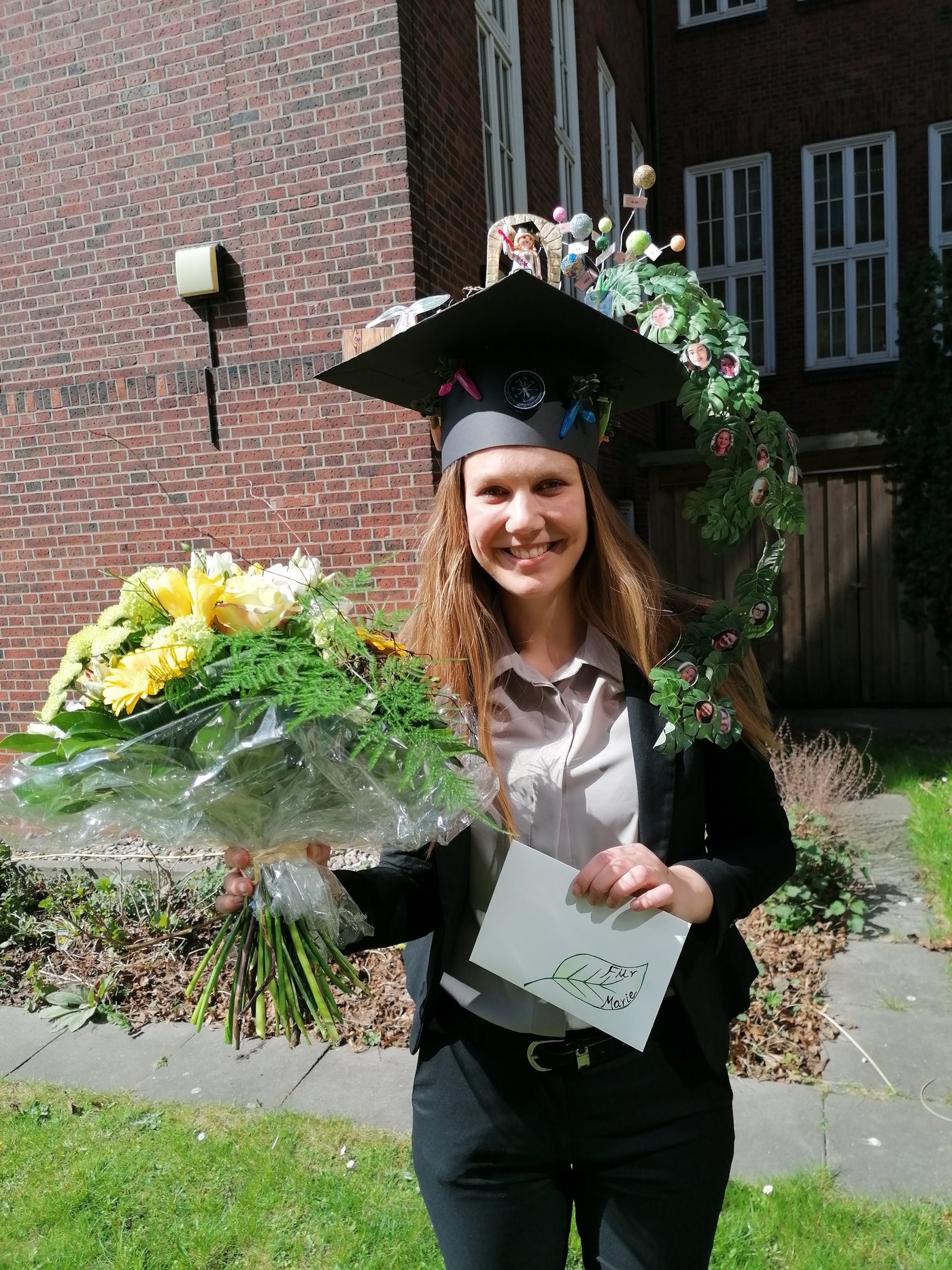Marie Groneberg kurz nach der Dissertation. Sie trägt einen selbstgebastelten, bunten Doktorhut auf dem Kopf, in der rechten Hand einen großen Blumenstrauß und in der linken Hand ein Urkunde.
