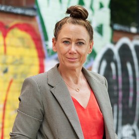 Dr. Meike Pahlmann: a woman with a light brown costume jacket and red jumper underneath (with V-neck and chain). She wears a bun on her head and stands in front of a graffiti wall.