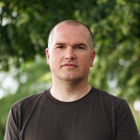 Dr Jan Priebe: a researcher with very short hair and a brown shirt stands outside in front of a leafy background.