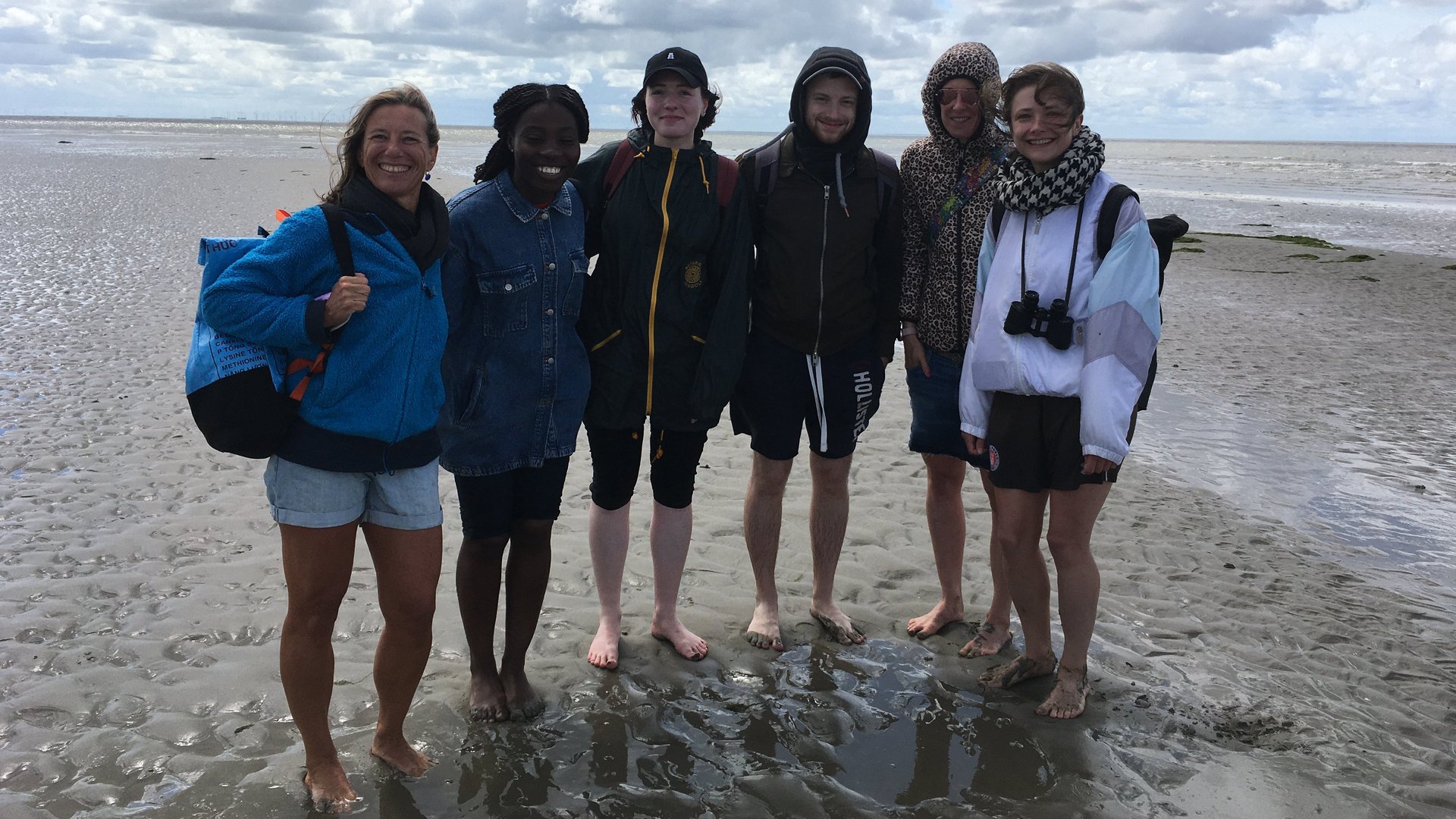 AG Helminthen-Immunologie im Watt bei Büsum: eine Gruppe von 6 Menschen stehen barfuß und mich hochgekrempelten Hosen im Watt und lachen in die Kamera. Im Hintergrund ist ein wolkiger Himmel zu sehen.