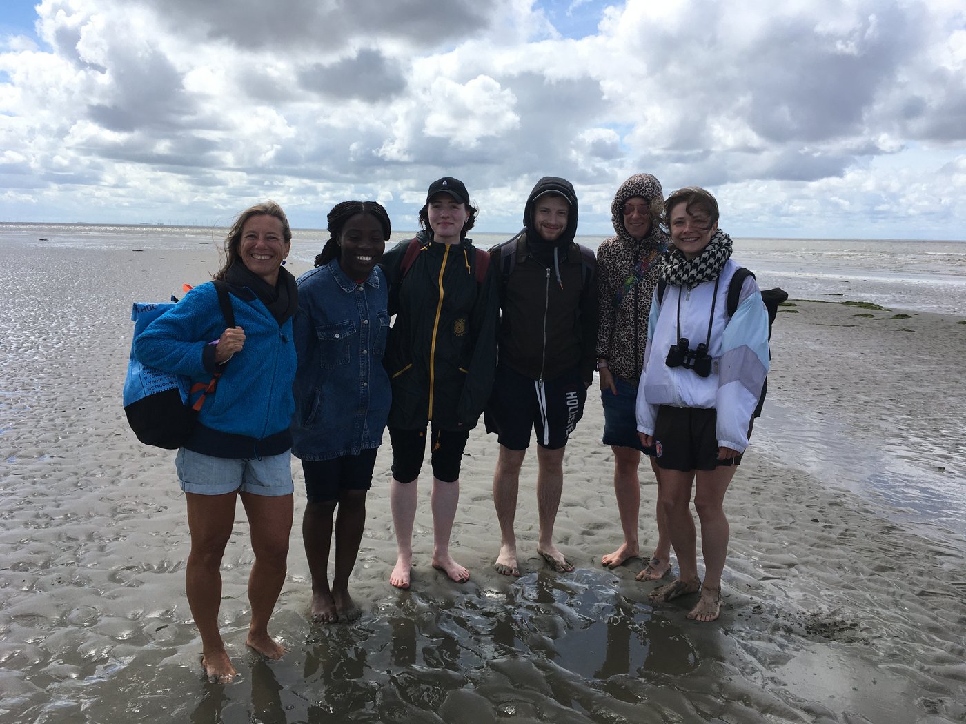 AG Helminthen-Immunologie im Watt bei Büsum: eine Gruppe von 6 Menschen stehen barfuß und mich hochgekrempelten Hosen im Watt und lachen ausgelassen in die Kamera. Im Hintergrund ist ein wolkiger Himmel zu sehen.