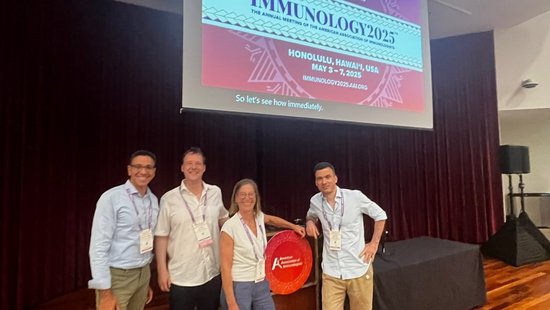 Presenting at the annual meeting of the American Association of Immunologists Four people stand smiling in front of a sign and large screen displaying “Immunology 2025” at a conference.