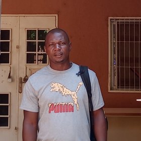 Kékoura Ifono: a man in a light-coloured Puma T-shirt. He is standing in front of a house entrance with a white lattice gate that leads into a reddish-brown courtyard.