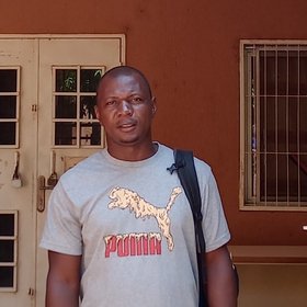 Kékoura Ifono: ein Mann in hellem Puma T-shirt. Er steht vor einem Hauseingang mit weißem Gittertor, das in einen rotbraunem Hof führt.