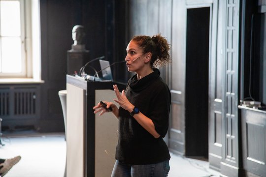 Auf dem Foto ist eine Frau zu sehen, die im dunklen Hörsaal des BNITM steht und einen Vortrag hält. Sie hat braune Haare, trägt einen Pferdeschwanz und gestikuliert mit den Armen.