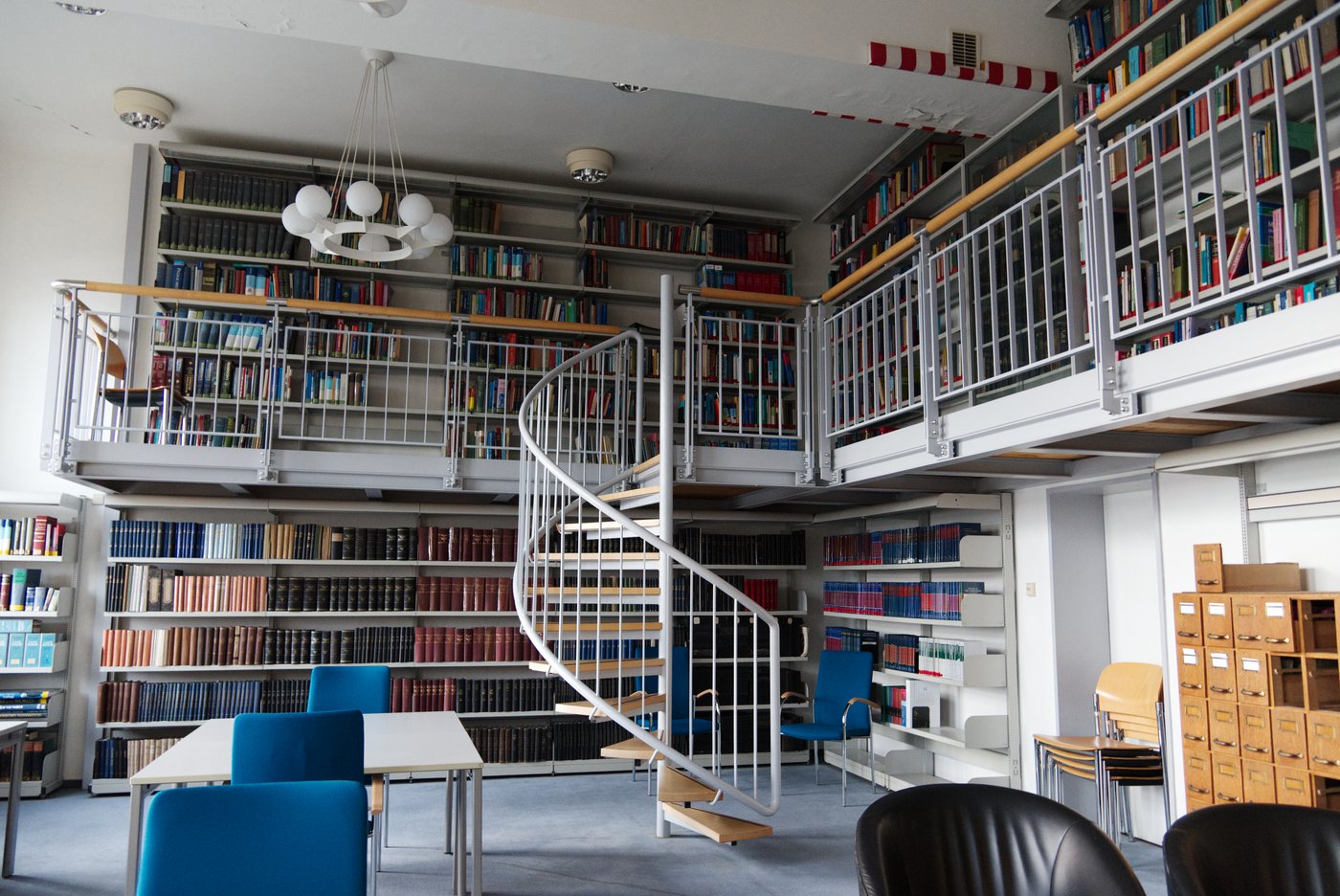 Zu sehen ist die Bibliothek des BNITMS, ein großer, heller Raum mit hohen Decken, in dem eine zweite Eben eingezogen wurde, die man über einekmetallende Wendeltreppe erreicht. An den Wänden stehen Bücherregale mit Büchern. In der Mitte des Raumes stehen kleine Tische mit blauen Stühlen.