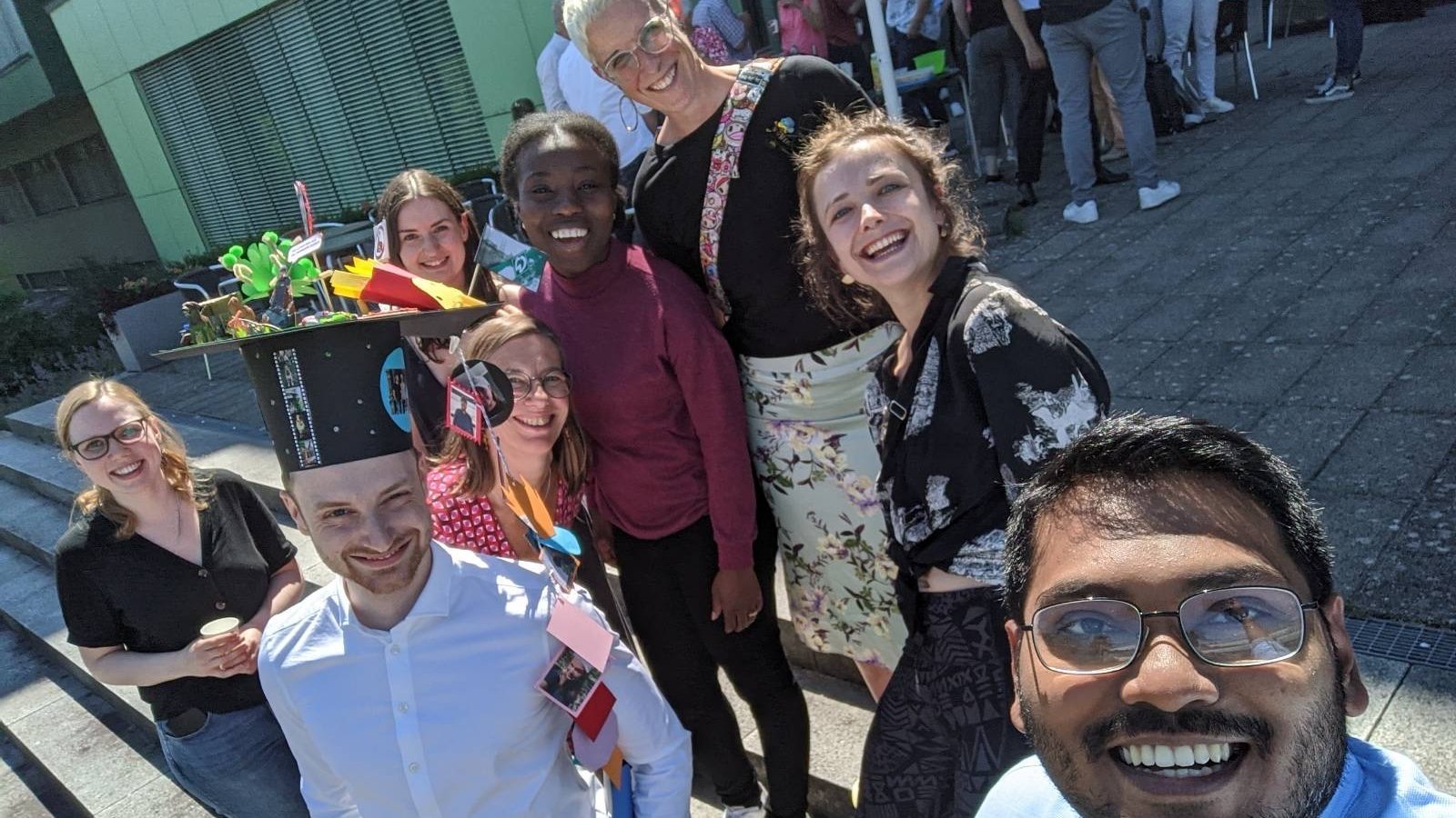 Dissertationsfeier Lennart Heepmann: zu sehen ist die AG Helminthen-Immunologie, die auf einer Treppe in der Sonne ein Selfie macht. Eine Person trägt einen selbstgebastelten Doktorhut, der Rest der Gruppe steht lachend um Ihn herum. Alle schauen in die Kamera.