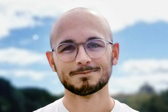 Louis Sarrazin Louis Sarrazin: Ein Doktorand steht lacht freundlich in die Kamera. Er trägt eine Glatze, einen schwarzen Kinn- und Oberlippenbart, ein weißes Shirt und eine runde Brille. Er steht draußen vor einem blauen, aber leicht wolkigem Himmel.