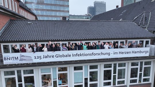 Drohnenbild des BNITM vom Hafen aus: Mitarbeitende stehen auf dem Balkon und winken. Darunter ein Banner mit der Aufschrift "125 Jahre Infektionsforschung - im Herzen Hamburgs"