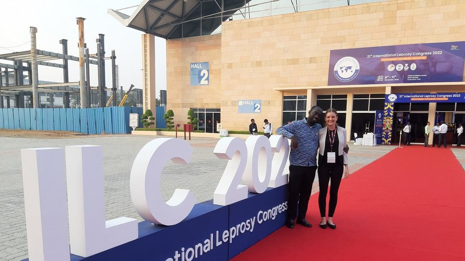 Team Groger Lead und Programmmanager des ghanaischen nationalen Leprakontrollprogramms auf der ILC 2022 in Indien: zwei Teilnehmer:innen stehen vor einem großen Sandsteingebäude, ein roter Teppich führt zum Eingang. Links der Beiden ist eine Schriftzug ILC 2022, 21st International Leprosy Congress zu sehen. Die beiden lächeln in die Kamera.