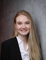 Eine Studentin mit langen, offenem, blonden Haaren lächelt in die Kamera. Sie trägt eine weiße Bluse und darüber einen schwarzen Blazer.