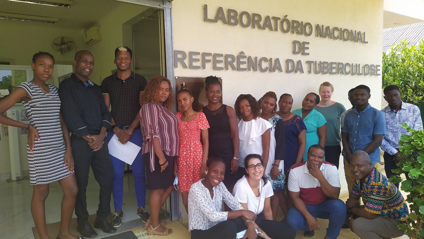 Staff of the LNR-TB in São Tomé  Das Foto zeigt sechzehn Sao Tome und Principe Mitarbeiter:innen des Laboratório Nacional de Referência da Tuberculose zusammen mit zwei Mitarbeitern des BNITM vor dem Laborgebäude in São Tomé und Príncipe. Die Gruppe steht vor einer Hasuwand, auf der der Schriftzug "Laboratório Nacional de Referência da Tuberculose" zu sehen ist.