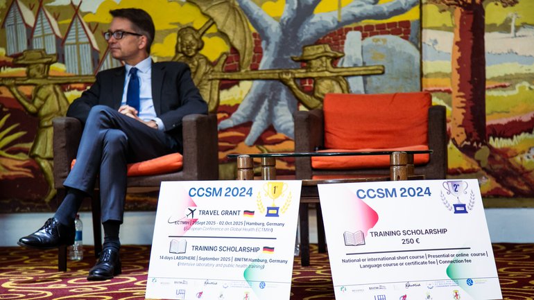 A seated man in a suit beside displayed CCSM 2024 award boards for travel grants and training scholarships, with a colorful mural in the background.
