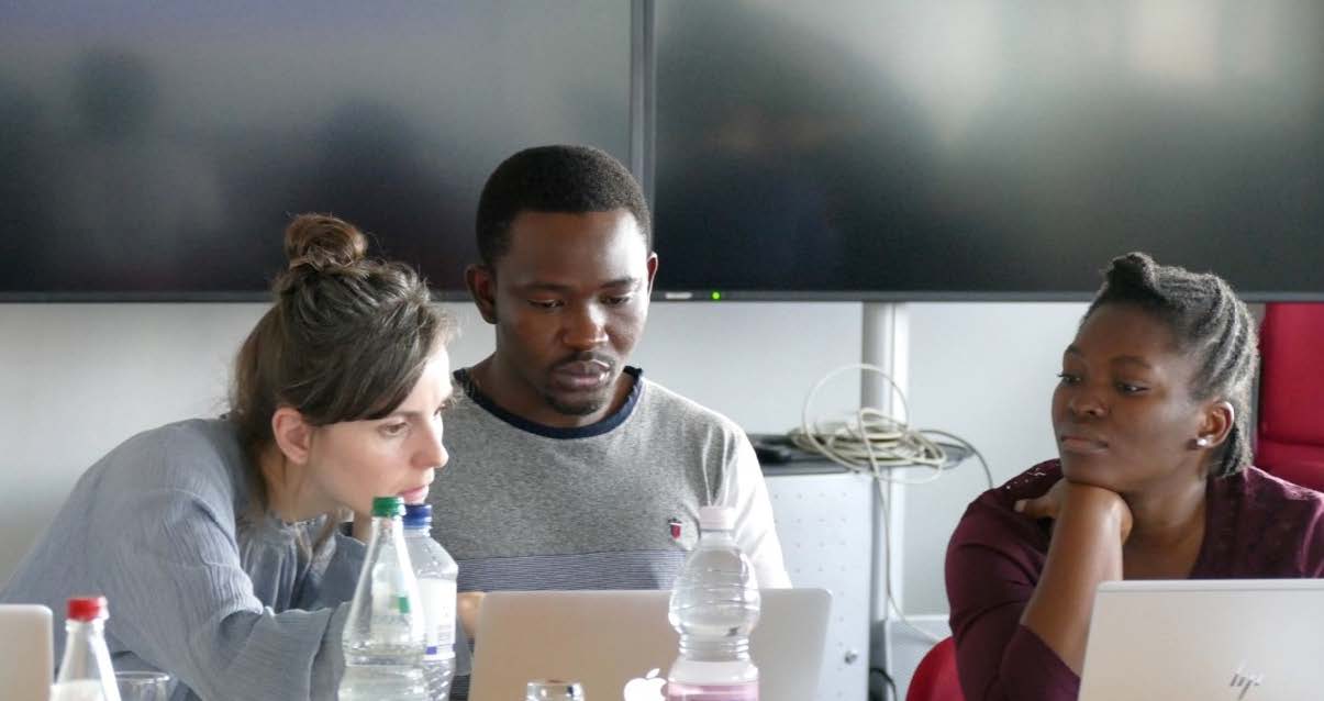 Das Foto zeigt drei diverse Menschen im Kursussaal des BNITM vor einer Lehrtafel am Laptop sitzen und diskutieren.