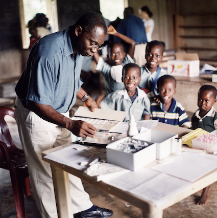 Ein Wissenschaftler des KCCR sieht sich Malariablutausstriche auf Objektträger (Glasplatten) an. Im Hintergrund sind Kinder zu sehen, die lachen.