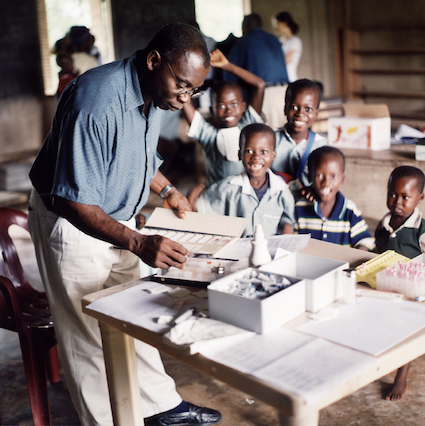 A KCCR scientist looks at malaria blood smears on microscope slides (glass plates). Children can be seen laughing in the background.