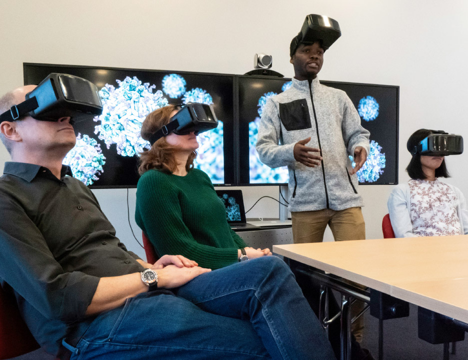 Mehrere Personen mit VR Brillen. Ein Mann spricht mit hochgeschobener Brille in die Runde