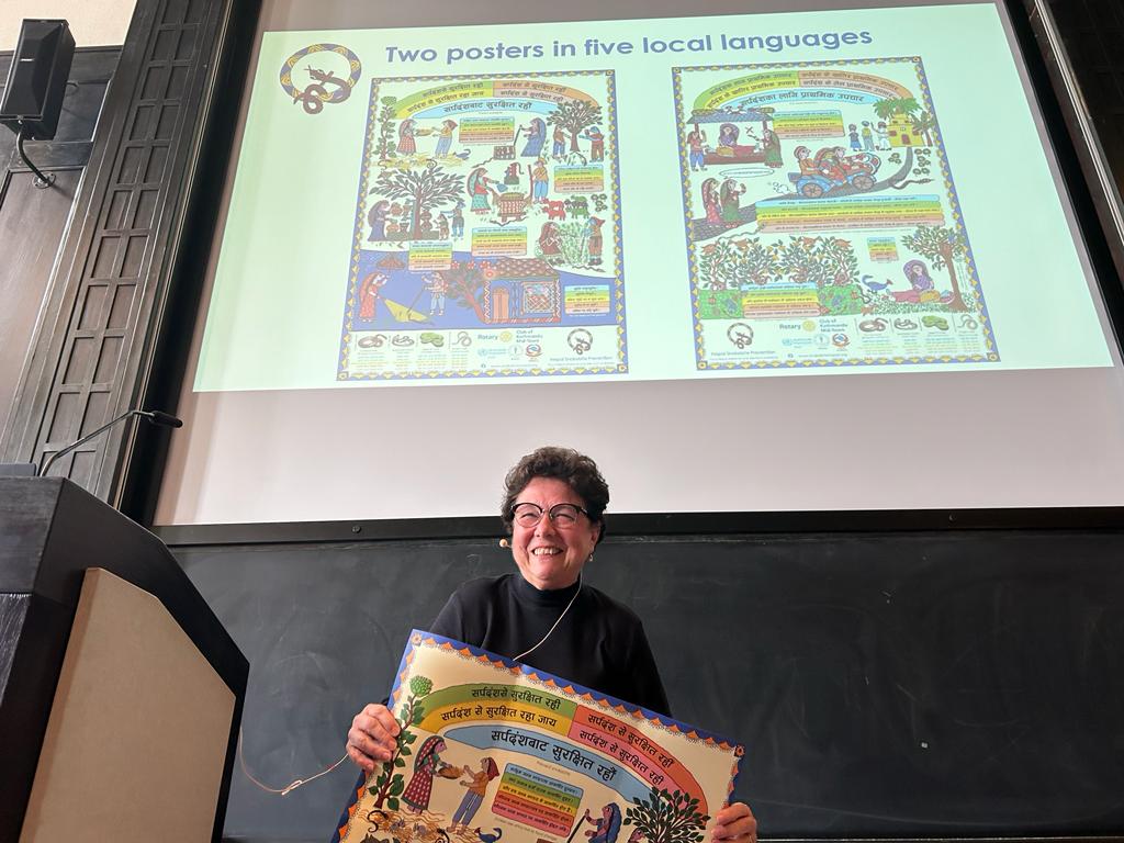 The picture shows a lecture holding a snakebite community poster into the camera. In the background both sides of the poster can be seen projected.