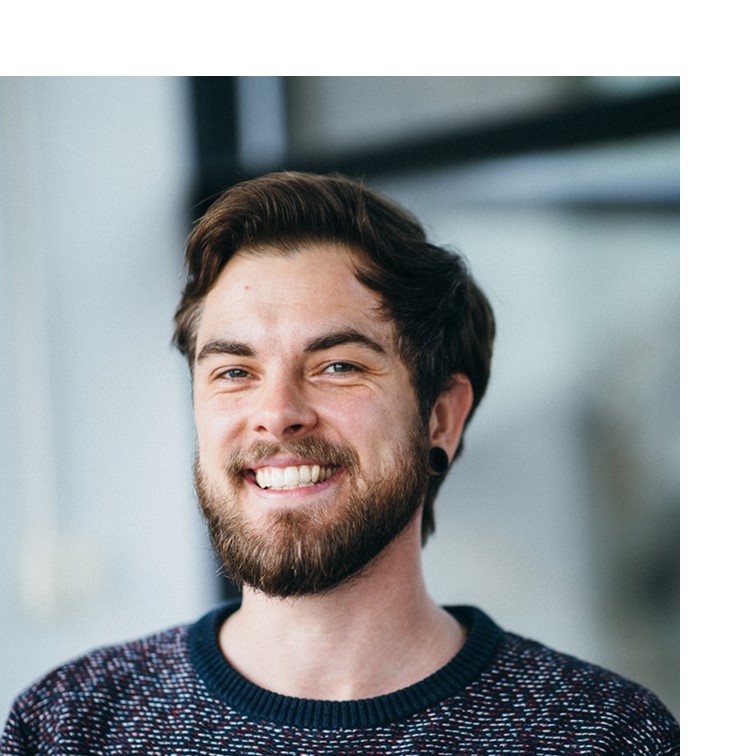 [Translate to English:] Portrait of a white male bearded person, wearing a blue-red-white sweater, smiling into the camera.