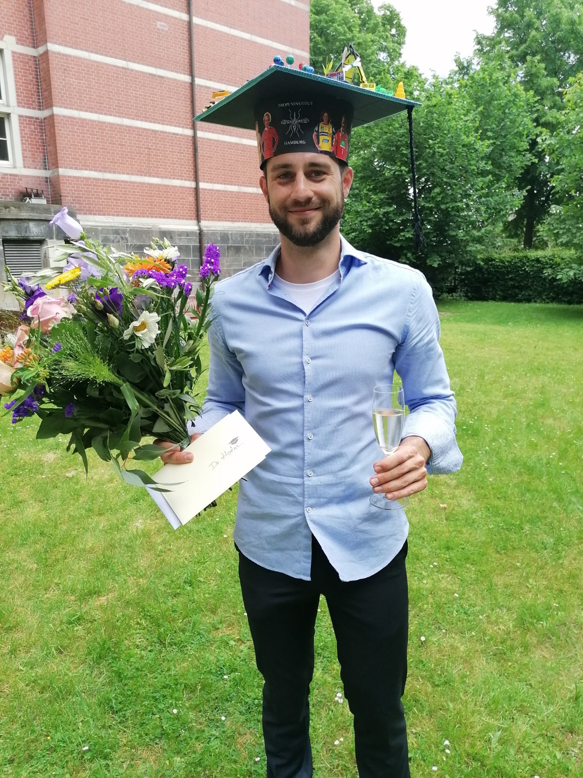 Stefan Hoenow kurz nach seiner Dissertation. Er trägt einen selbstgebastelten, bunten Doktorhut auf dem Kopf, in der rechten Hand einen großen Blumenstrauß und in der linken Hand ein Sektglas.
