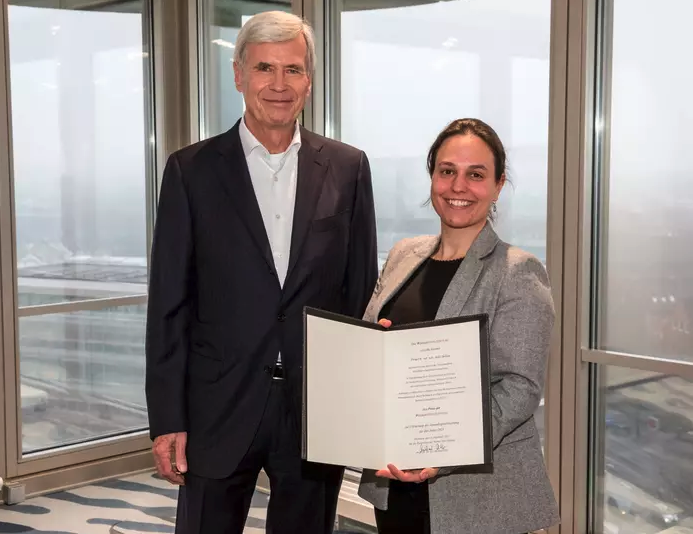 Dr. Julie Sellau erhält in einem festlichen Rahmen die Urkunde überreicht. Im Hintergrund sieht man bodentiefe Fenster und  einen grauen Wintertag.