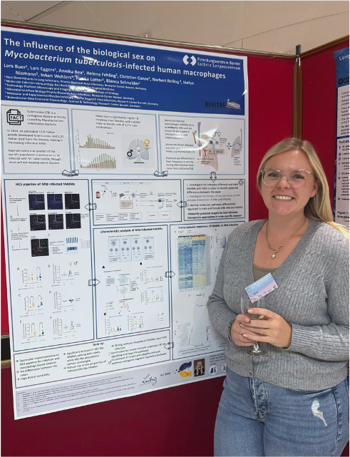 Lara Buer stands to the right of her poster and smiles warmly at the camera. She is wearing jeans and a gray pullover.