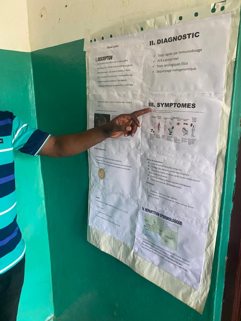 Poster presentation on Lassa virus at LFHV-GKD. Guéckédou, Guinea, August 2025 One laboratory member is pointing to a poster on Lassa fever which is hanging on the wall
