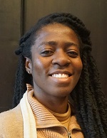 Eine Forscherin mit langem schwarzen Haar, das zu kleinen Zöpfen geflochten ist, lächelt in die Kamera. Sie trägt ein gelbes Oberteil und steht vor einer dunklen Wand.