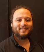 Ein Clinical Data Manager mit Bart, langem Haar, das zu einem Zopf gebunden ist und einem schwarzen Hemd steht vor einem dunklen Hintergrund.