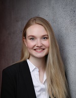 Eine Studentin mit langen, offenem, blonden Haaren lächelt in die Kamera. Sie trägt eine weiße Bluse und darüber einen schwarzen Blazer.