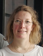 Eine medizinische Studentin mit kinnlangem, blonden Haaren, einer runden Brille mit goldenem Rand, eine goldene Kette und einem weißes Tshirt steht vor einem gläsernden Durchgang.